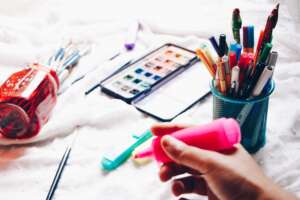 person holding pink highlighter with pens and paint brushes in the background