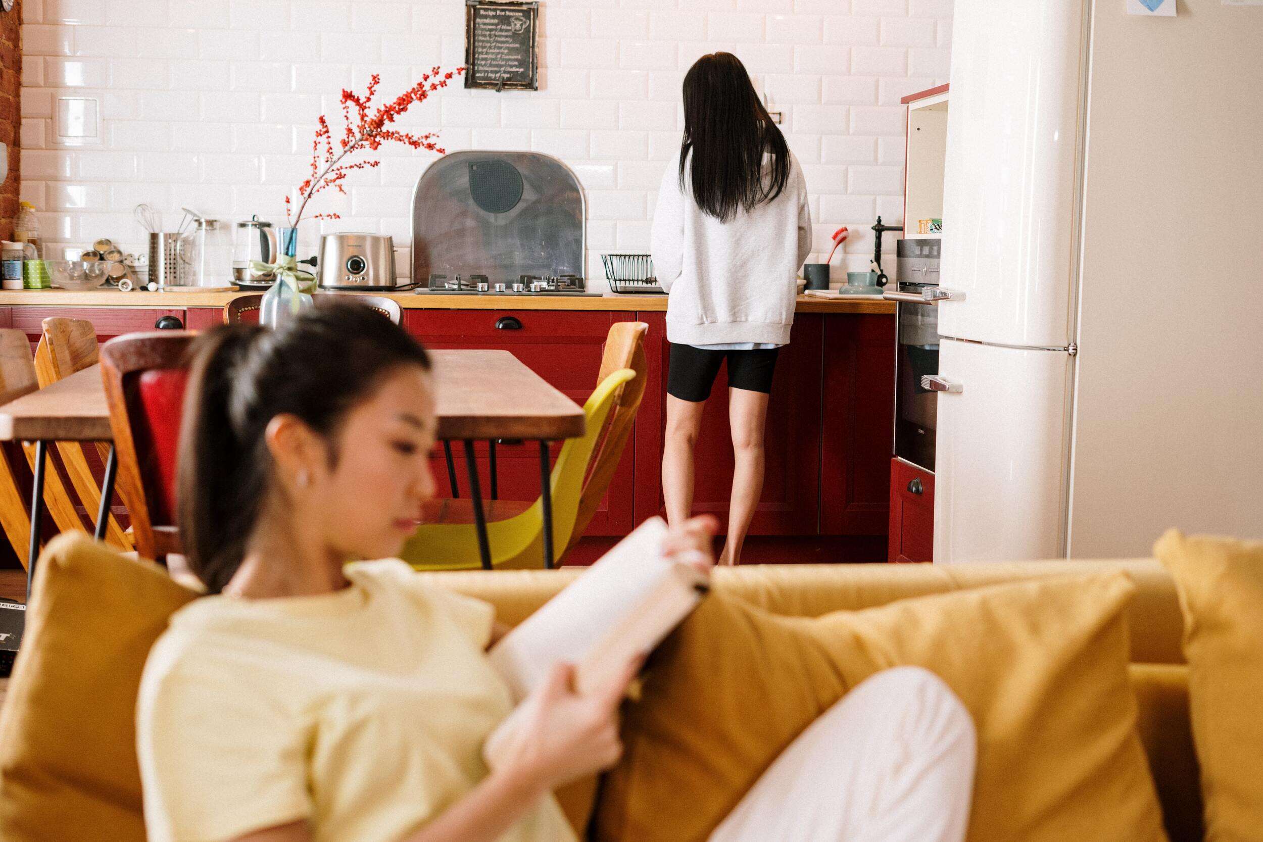 women sitting on a yellow couch reading a book and another women standing behind her