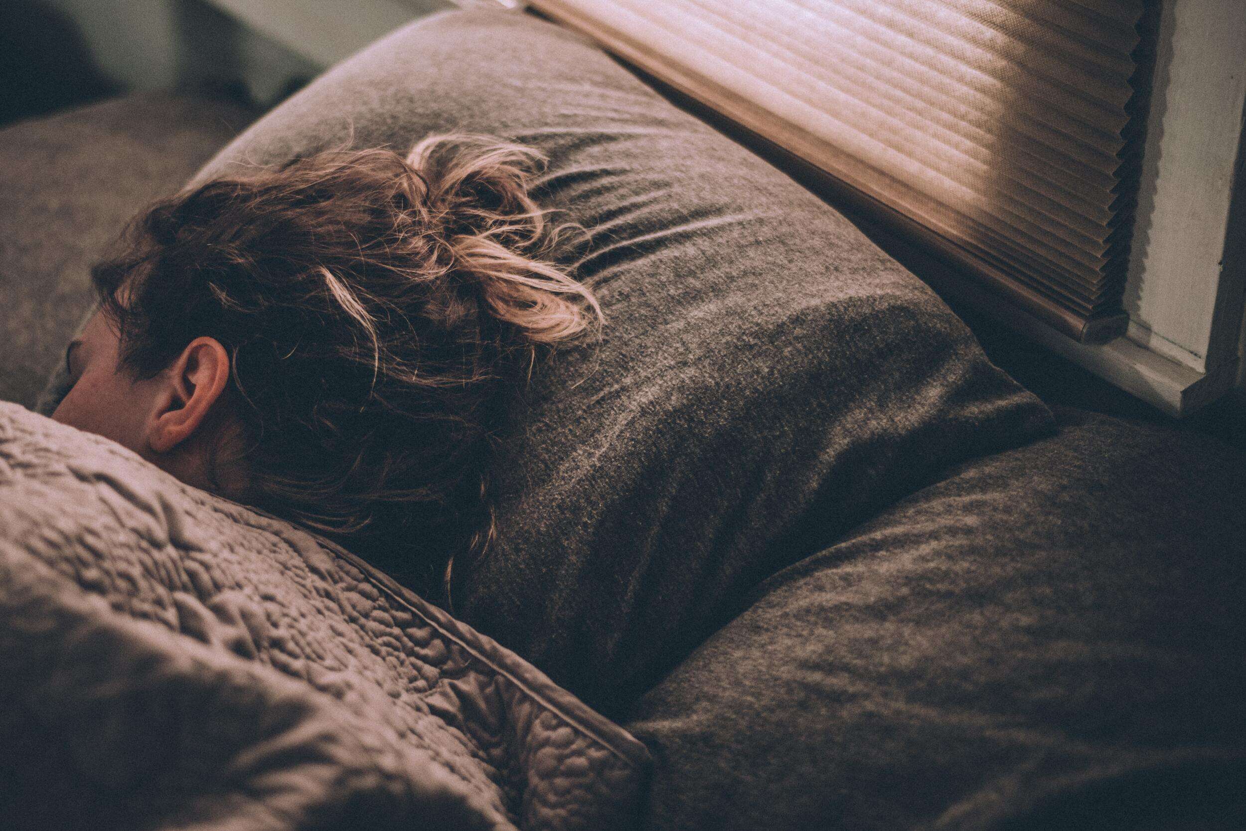18 Tips for Sleeping Better in College women sleeping in bed under dark gray blanket