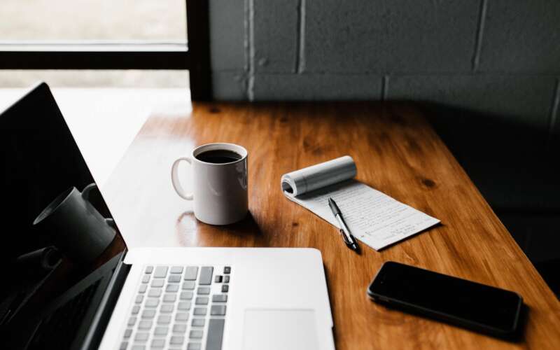 MacBook Pro, white coffee cup, notepad, and cell phone sitting on a wood table beside a wall
