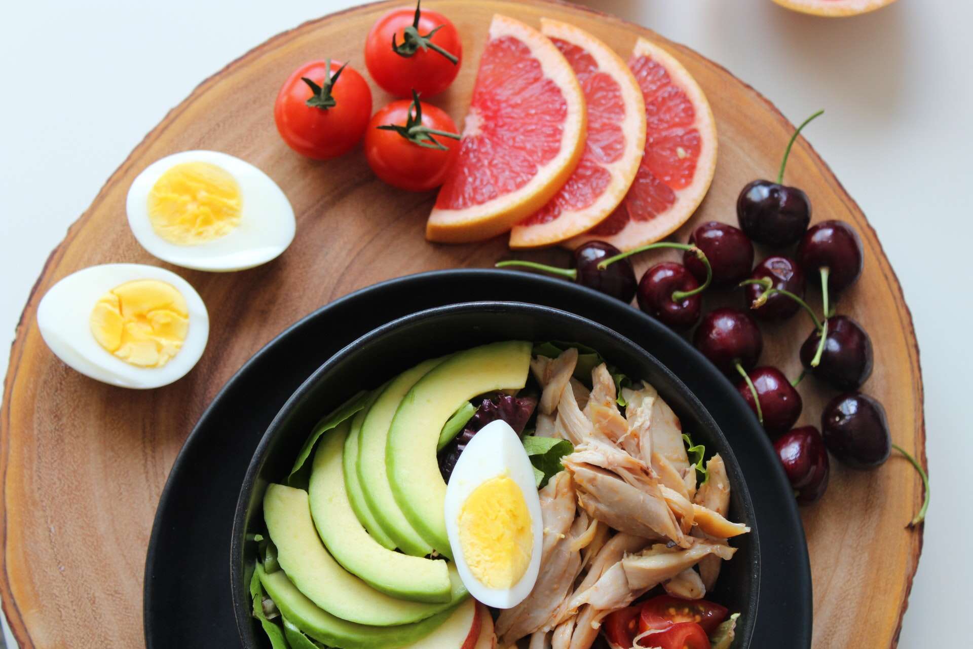 A bowl filled with slices of avocado, a hard boiled egg, and pieces of chicken with other healthy foods around the outside of the bowl