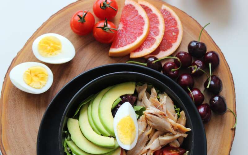 A bowl filled with slices of avocado, a hard boiled egg, and pieces of chicken with other healthy foods around the outside of the bowl