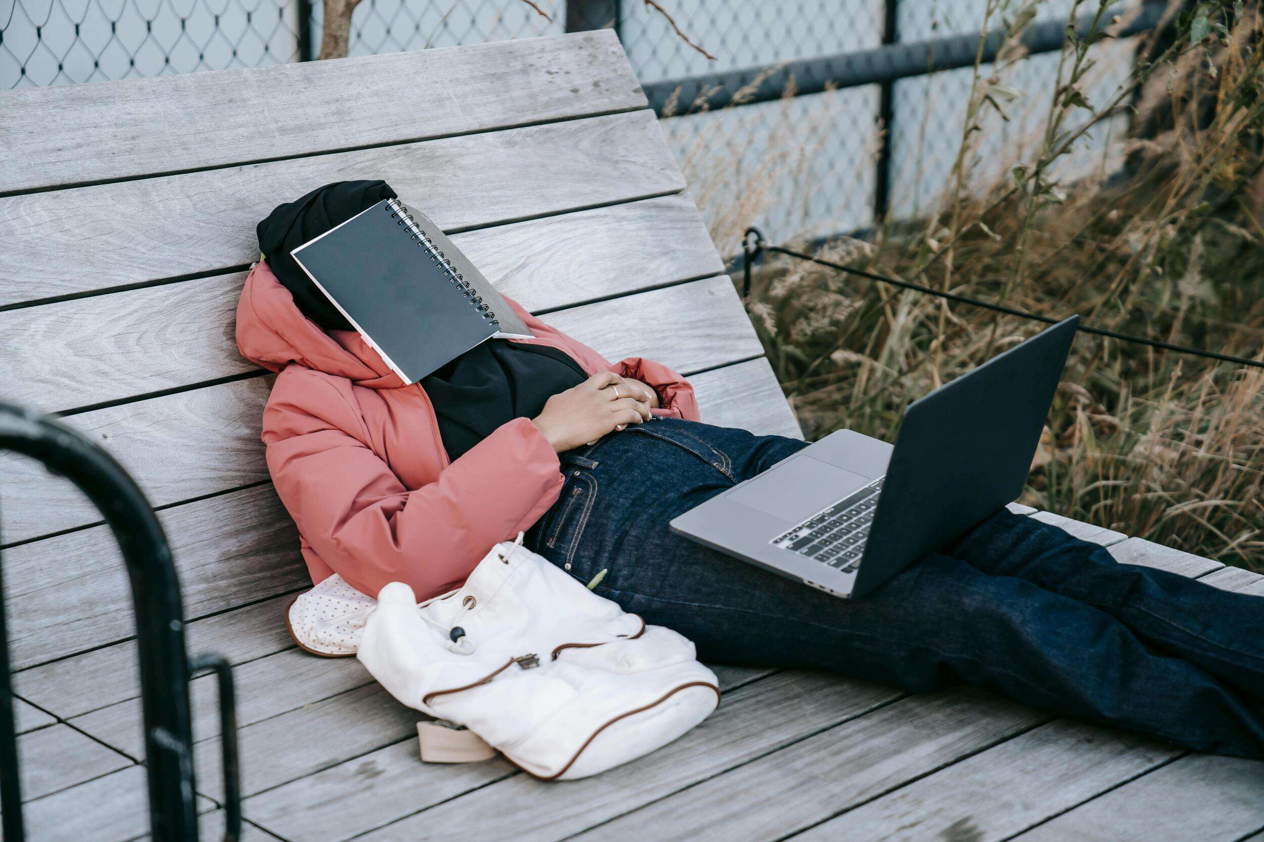 college student sleeping on a bench outside with a notebook over his face