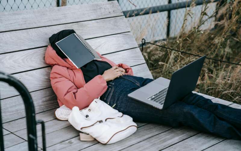 college student sleeping on a bench outside with a notebook over his face