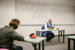 Student sitting at a desk taking a test while professor sits in front of them in a room with rules on whiteboard