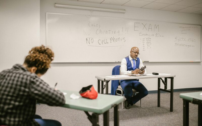 Student sitting at a desk taking a test while professor sits in front of them in a class room with rules on whiteboard