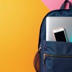 Dark blue college backpack up against a yellow wall with laptop, phone, and water bottle sticking out of it